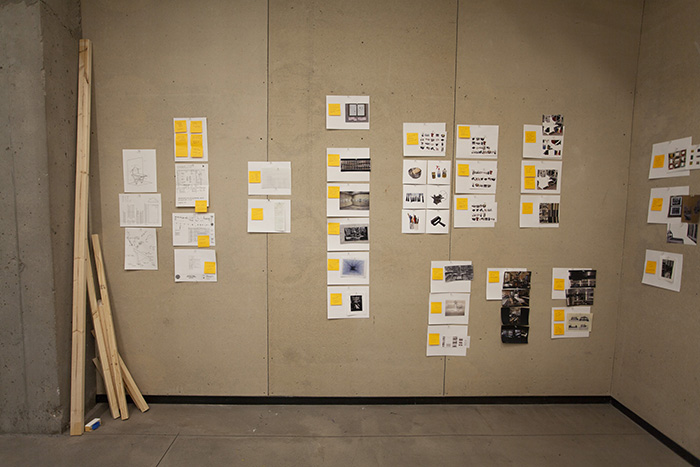 many small diagrams and prints, most with yellow post-its attached, arranged on a tan homasote wall. Sticks of wood lean in the left corner