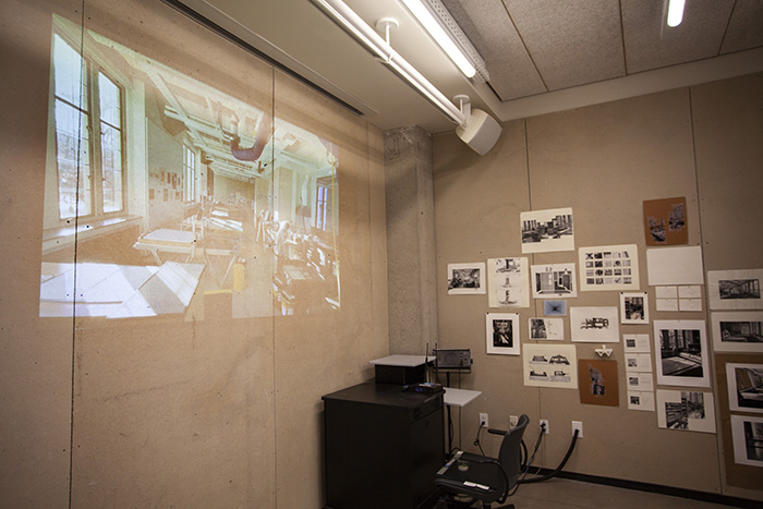 photo looking into a corner of a room with tan homasote walls; on the left wall is a video projection of a print studio; on the right wall is a salon-style arrangement of small photographs and drawings