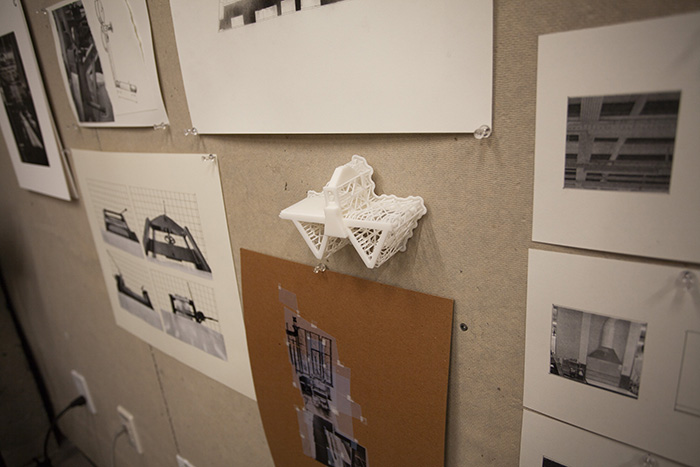 close-up on a wall with multiple diagrams and photos on it; in the center of the photo is a tiny white 3D printed printing press, with lots of ropey support structures still attached