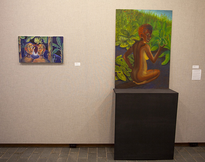 an installation on gray fabric walls of a small horizontal painting of two figures and a larger vertical painting of a single kneeling figure. The vertical painting is propped on a black pedestal.