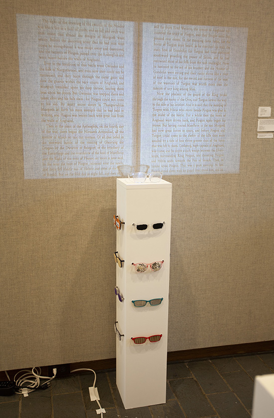 A white pedestal with fanciful glasses installed on it. A video is projected onto the wall just above and behind it, showing book pages and text. 