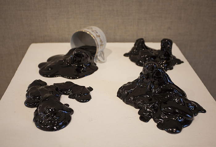 four blobs of shiny black goo on top of a white pedestal. The back left blob is emerging out of a teacup.