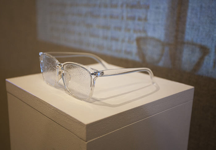 close up on a pair of glasses made of clear plastic with textured lenses sitting on top of a white pedestal; a video plays through them and casts their shadow on the video projection on the wall behind them