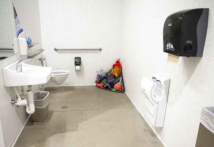 A bathroom with concrete floor and white tiled walls. Sink, mirror, trash can, toilet, and hand dryer are all visible. In the back right corner of the room is a pyramidal sculptural installation of a coral reef.