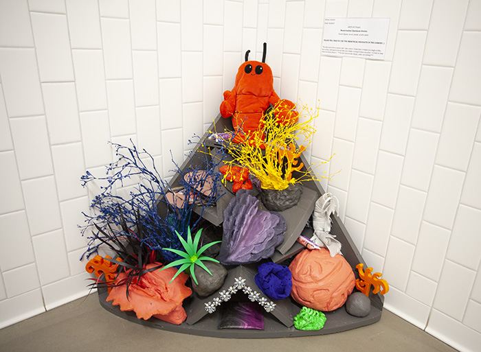 a sculpture of a pyramidal coral reef, with corals and animals, installed in the corner of a white-tiled space