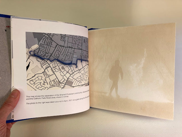 an open book spread. The left page is a map with the top half shaded in blue. The right page is a ghostly tan overlay of a figure with some sort of atmosphere, perhaps flames, in the background.