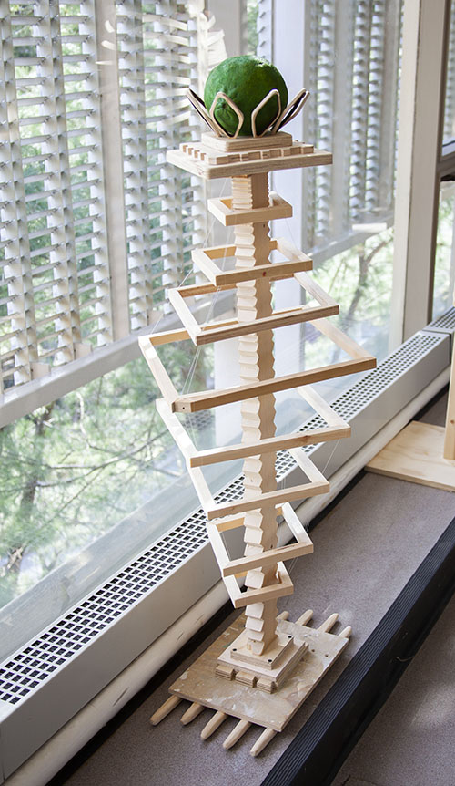 a wooden sculpture with square rings held in tension by fishing line around a central wooden spine. At the top is an oversized green line surrounded by petal-like wood structures.