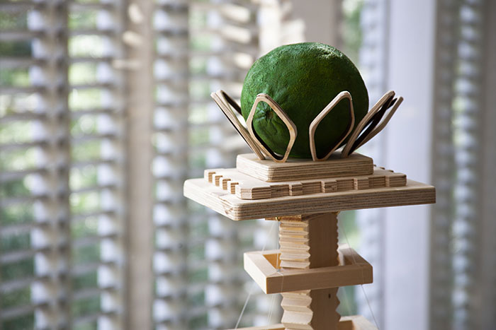 close-up of the top of a sculpture. A green oversized lime is balanced between wooden petal-like structures atop a layered wood platform