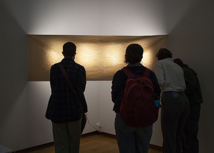 four people in silhouette standing in front of a pierced canvas, lit from behind, stretched across the corner of a gallery