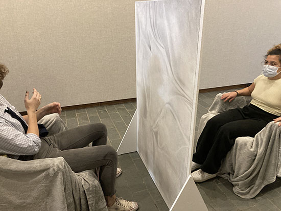 two people sit in blanket-covered chairs on either side of small freestanding wall. The side of the wall facing the viewer has a charcoal drawing of draped fabric on it