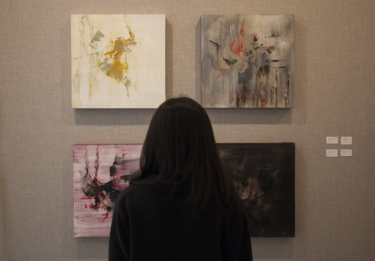 four abstract paintings on a gray fabric wall with a person in dark clothing, with long dark hair, standing in front of them, back to the camera