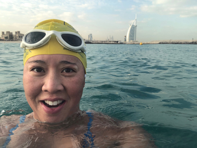 An alumna takes a selfie in the ocean with a city skyline behind.