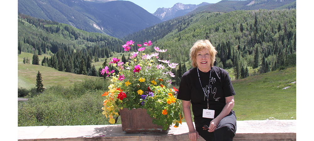Betsy sitting on a wall beside a flower pot in front of a mountainous landscape