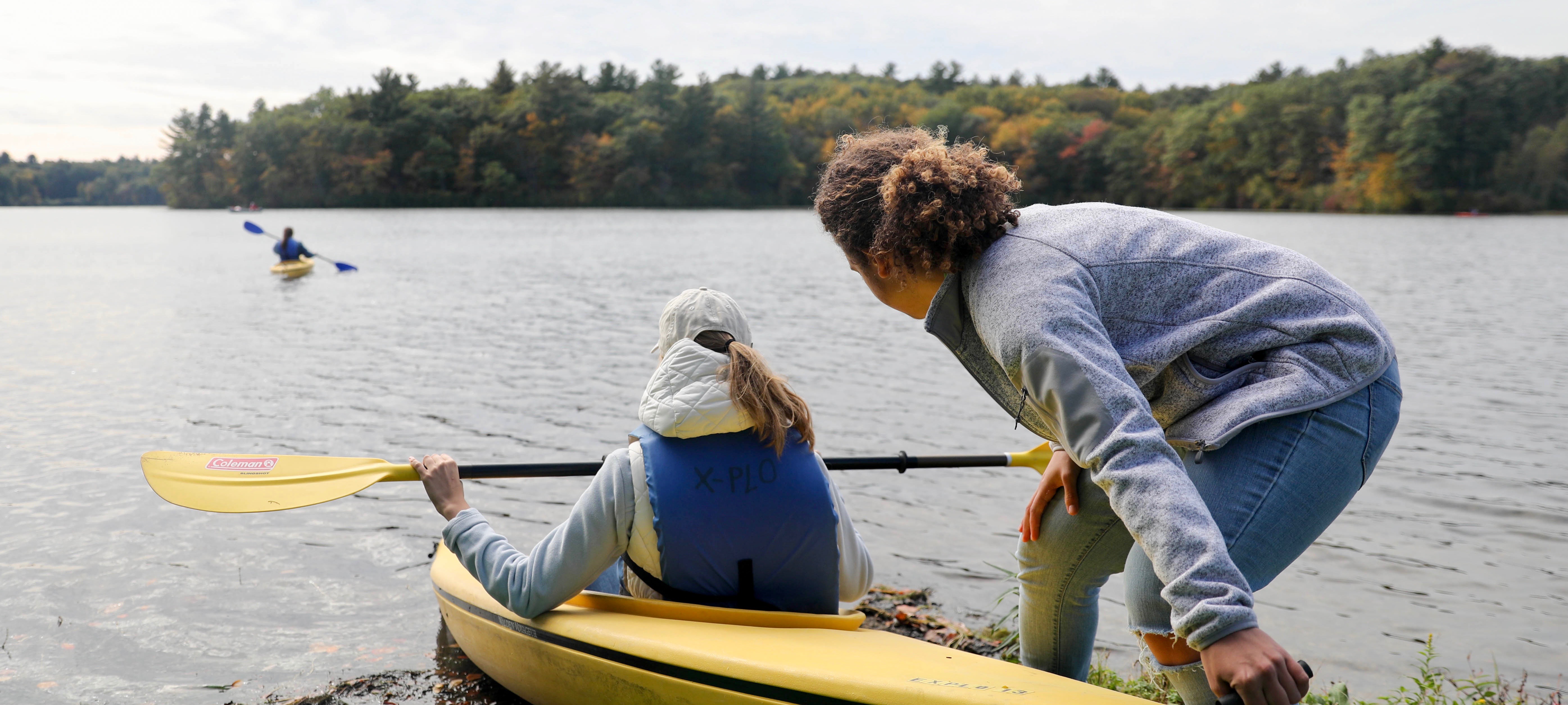A student sits in a yellow kayak, wearing a blue life jacket. Another student is pushing them off from the shallow water.