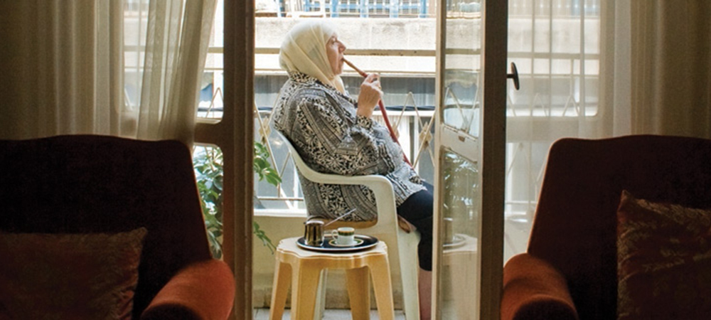 A still from the film Teta, Alf Marra. An old woman wearing a headscarf sits on an apartment balcony smoking from a hookah pipe.