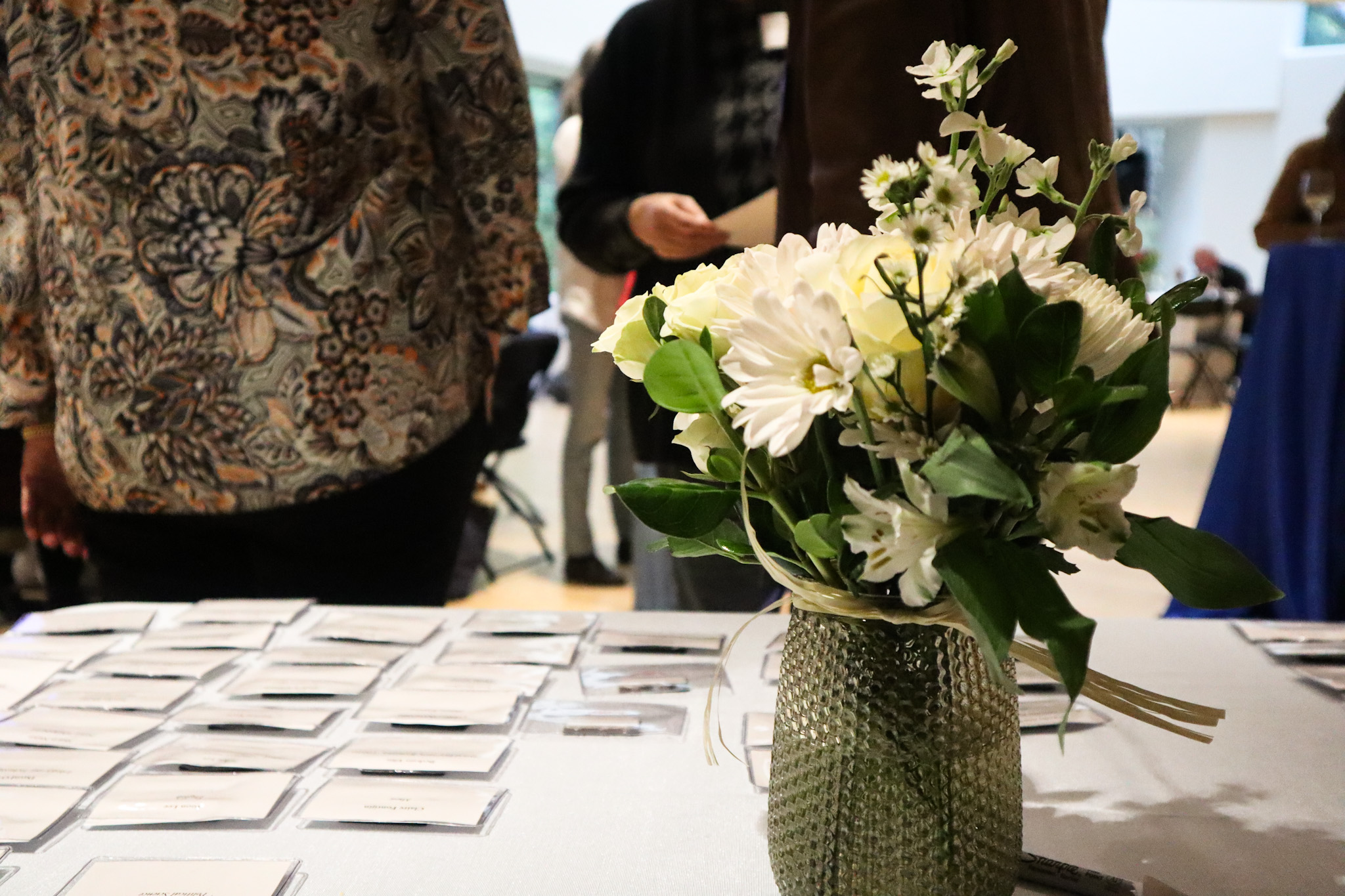 A table with nametags and a white bouquet of flowers in a vase. Employees are gathered in the background. 