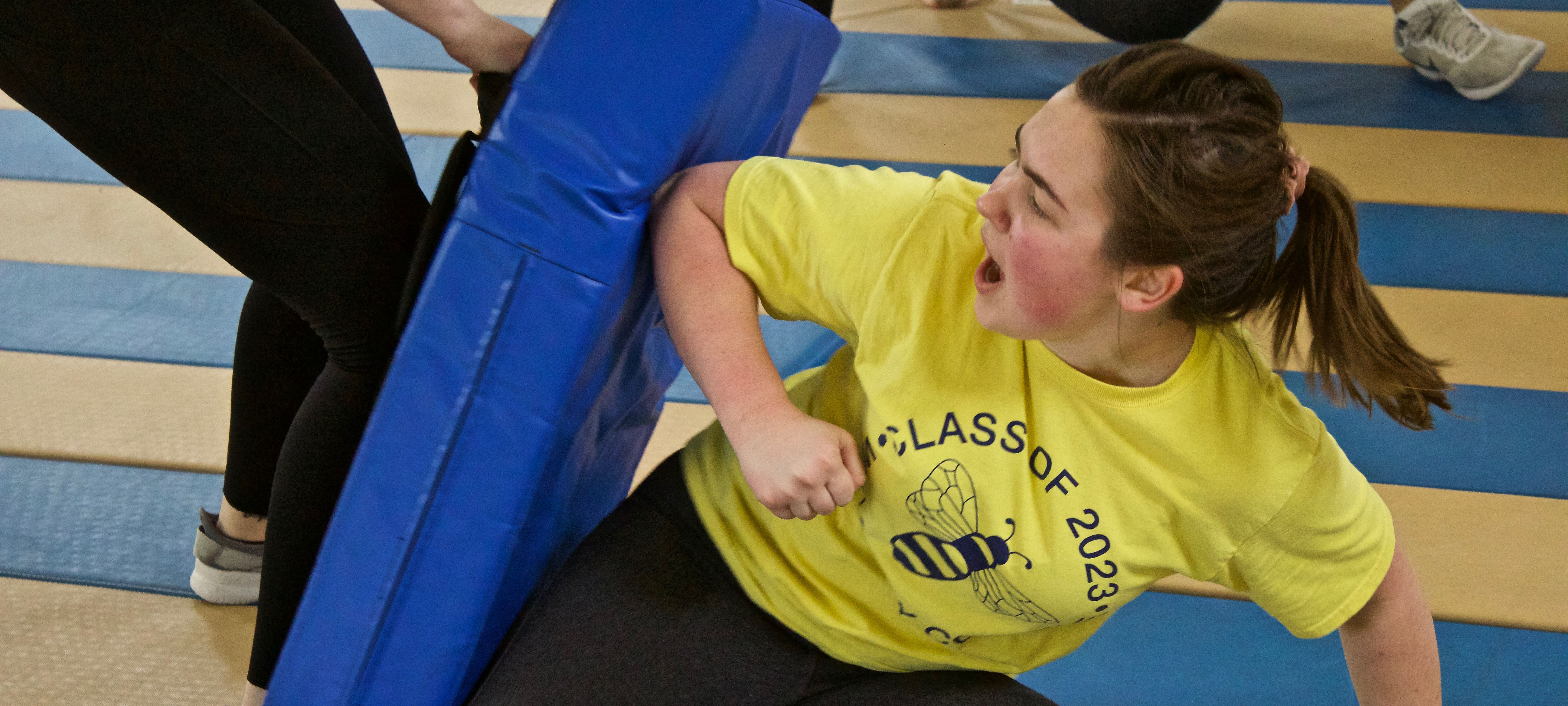 A student in a yellow Class of 2023 shirt is elbowing a blue, thick pad that another student is pushing onto them.
