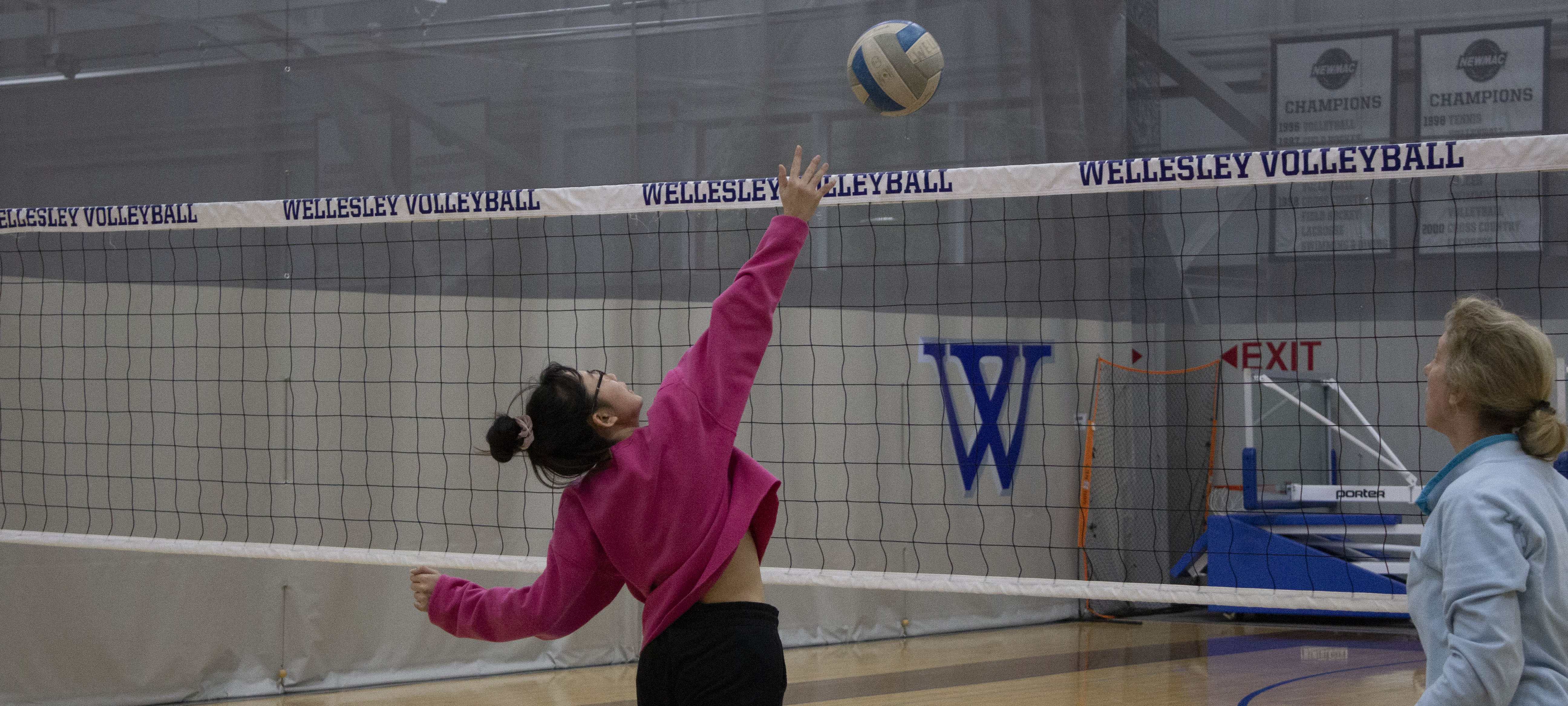 A student in front of a volleyball net with a centered blue W, throws a volleyball into the air to spike it. Their coach watches