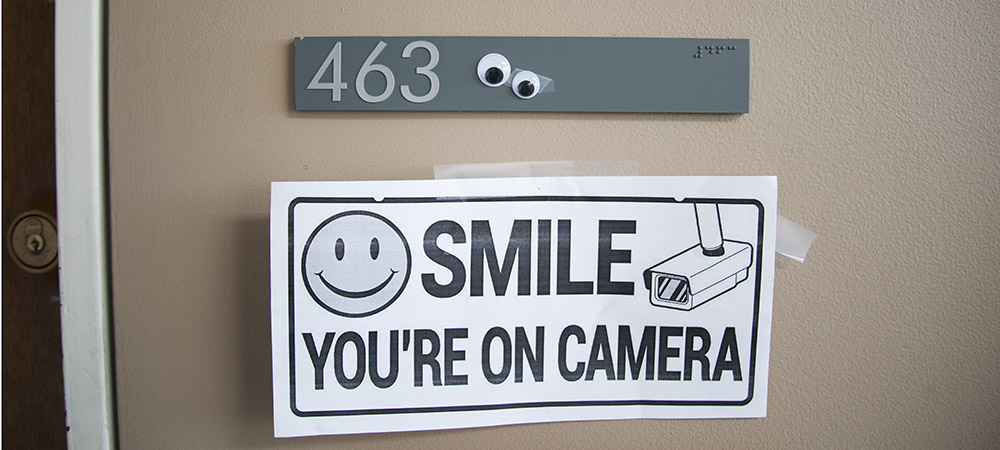 gray plaque with number 463 and two googly eyes on it, above white sign reading Smile, You're On Camera!