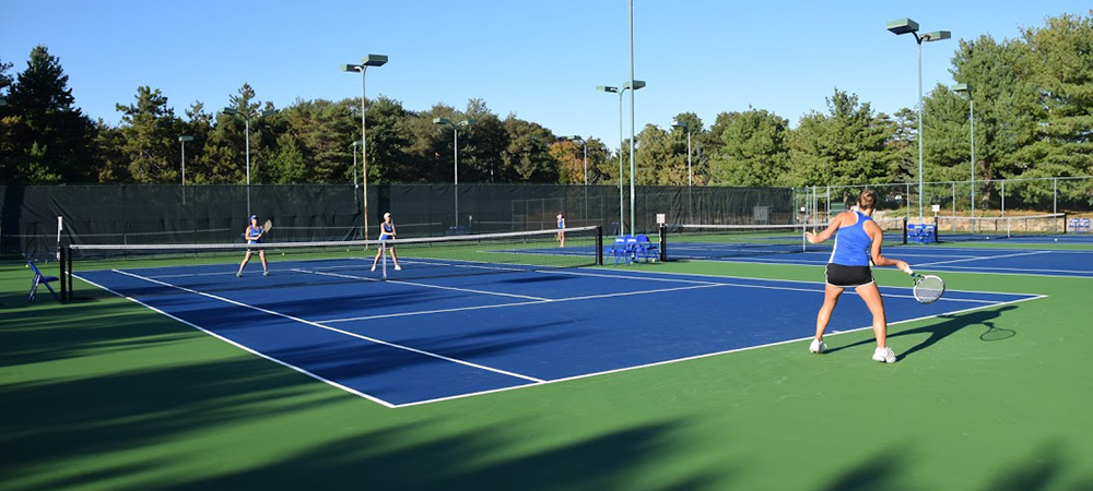 Three students are playing tennis on a hard court. Two are on the opposite side of the net from the other. 