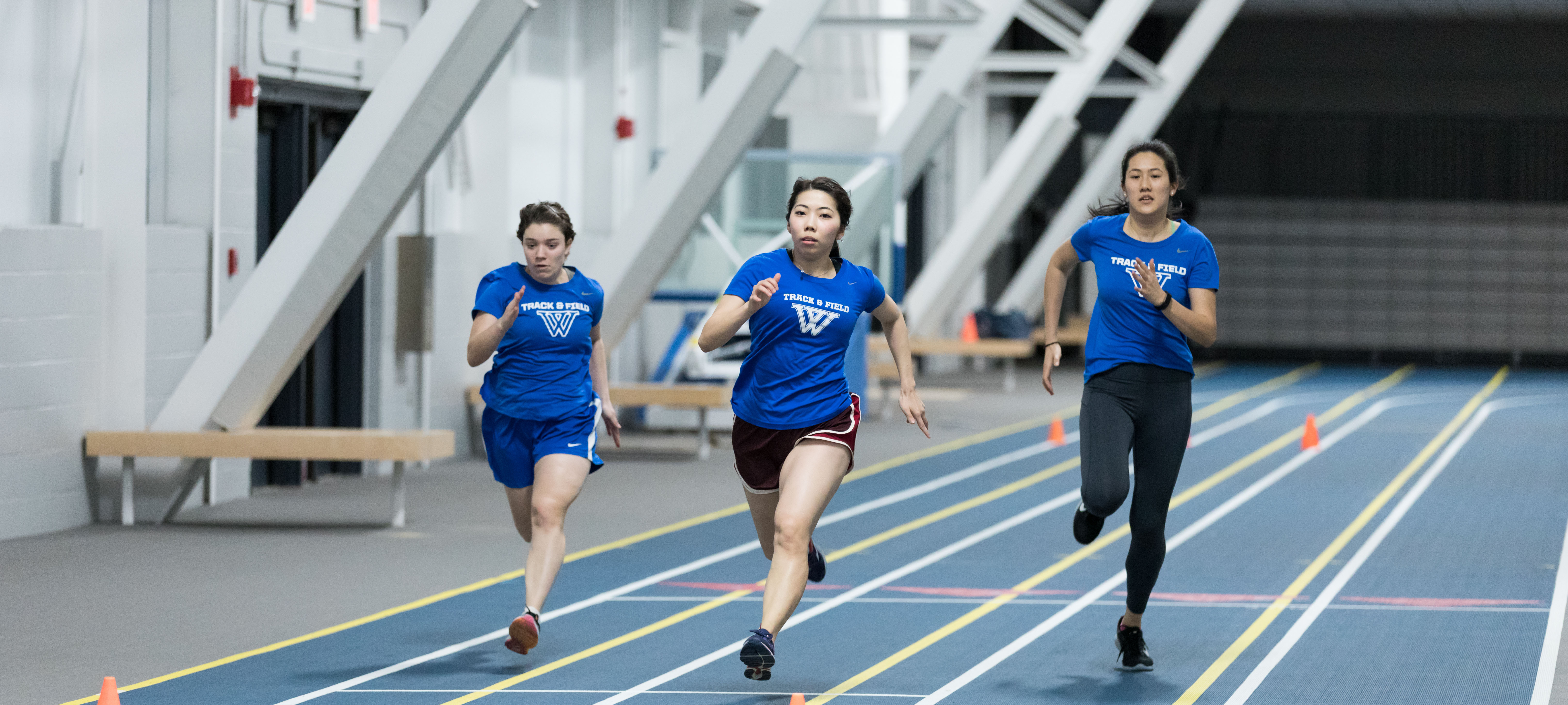 Three students in blue Wellesley Track & Field t-shirts are running down the KSC Track
