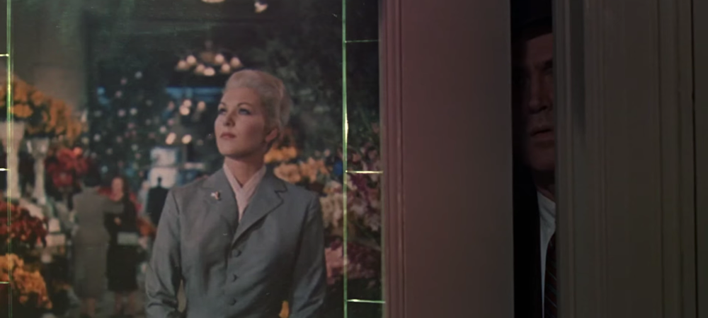 A still from the film Vertigo, a woman walks in a store and a man can be seen peering from an opening