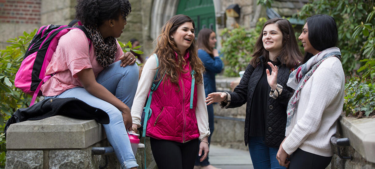 Picture of students on campus talking and laughing, surrounded by stone walls and buidings.