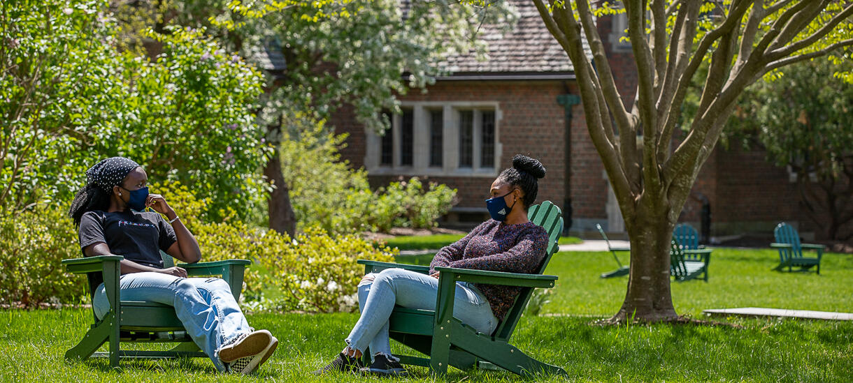 Picture of two students sitting outside, spring day, wearing masks, talking.