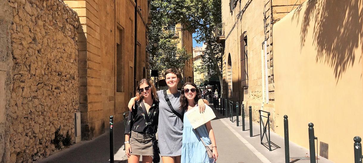 Three students stand together in the middle of a narrow street in Aix-en-Provence, France.