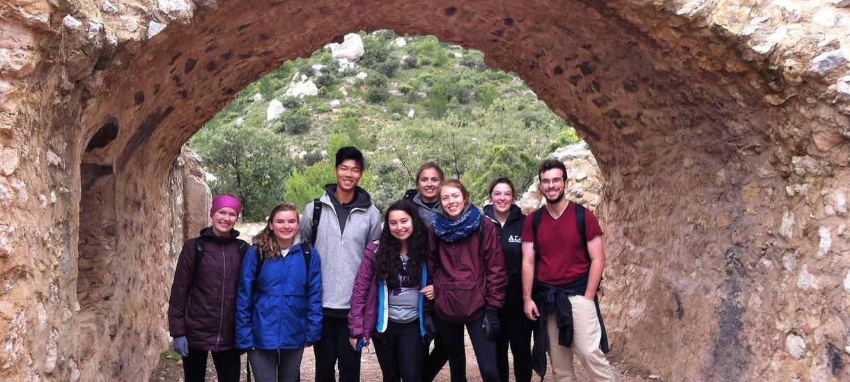 A group of eight students gathered for a photo underneath an arched rock structure, dressed in hiking gear.