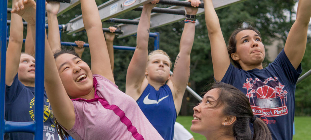 Student smiling in foreground with other students climbing monkey bar course