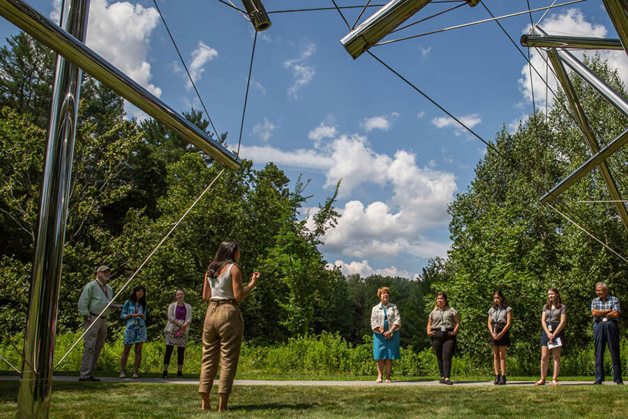 a group of people standing outside on grass while a student talks to them about an abstract metal campus sculpture