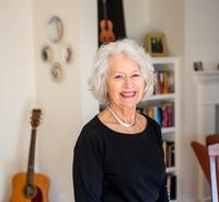 Lynne Viti wears a black top and sits atop a table, smiling at the camera; in the background a guitar and bookshelves can be see