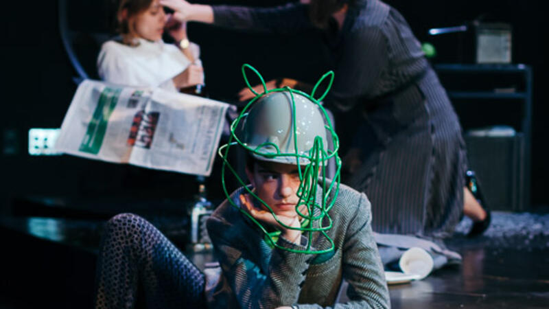 two actors on desk set in background behind actor in rhino head hat