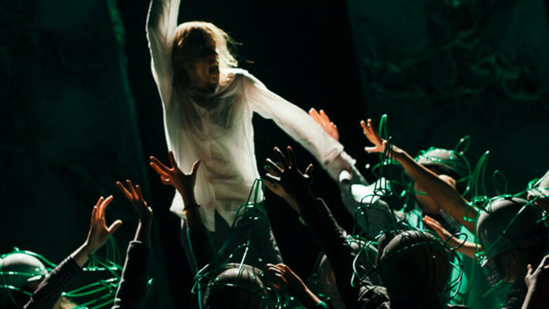 dimly lit stage with actor reaching up, surrounded by kneeling actors with rhino hats