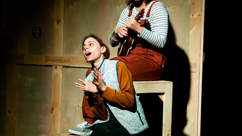two actors sitting on wooden structure, one playing ukelele