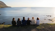 students, seen from behind, sit in a row looking out at sea