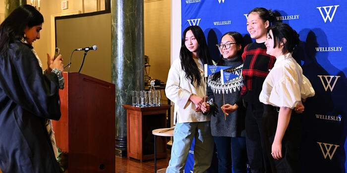 Four December graduates pose for a photo against a Wellesley logo backdrop. 