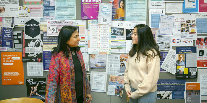 A student a guest speaker stand in front of a wall with posters