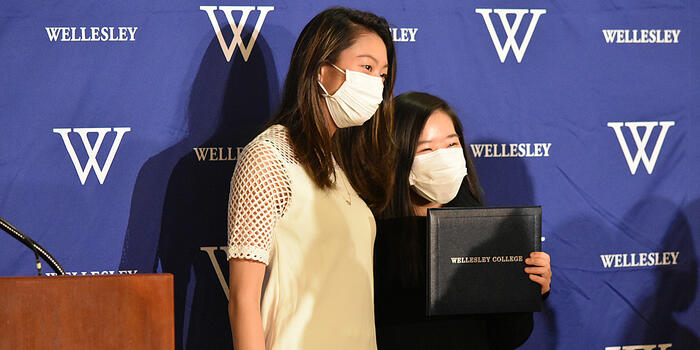 Two students pose for a photo with one student holding up her diploma case that read Wellesley College.