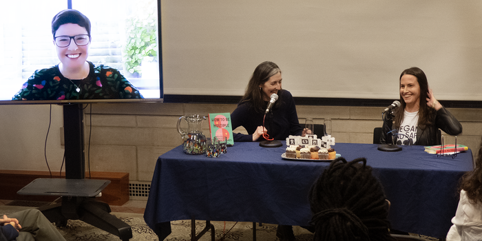 Three speakers at the front of a room including one on a video screen. All three are laughing. 