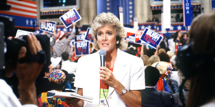 A woman reporter stands on the floor of a press room.