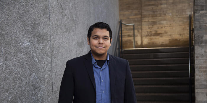 Man in blue shirt and black jacket stands in front of a building.