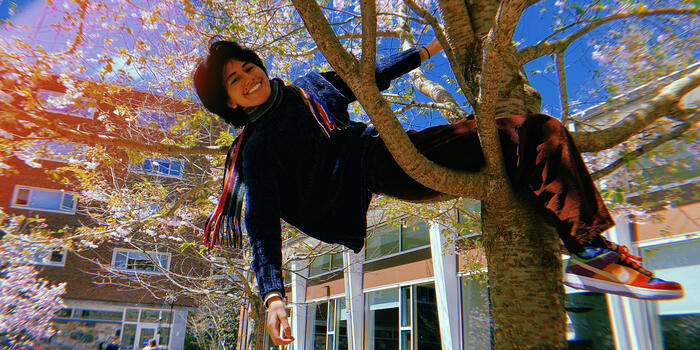 A student sits in the base of a tree with one hand grasping a branch and the other dangling behind her. She smiles at the camera
