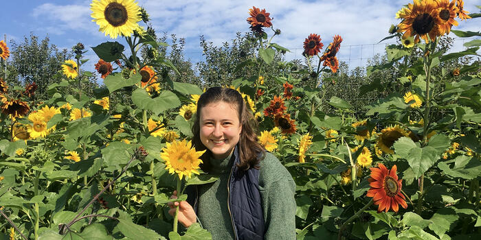Dominique Mickiewicz stands in a garden of sunflowers and smiles at the camera.