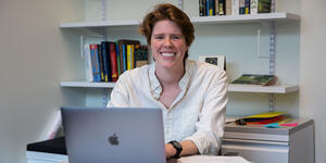New faculty member Erin Teich sits behind her computer and looks at the camera, smiling.