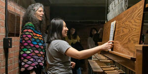 A student sits at the bench of a carillon with a teacher standing over her.