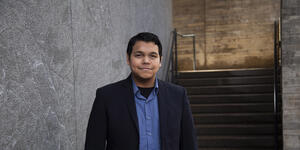 Man in blue shirt and black jacket stands in front of a building.