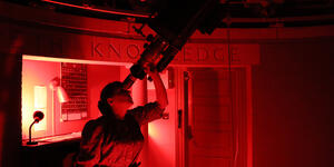 A woman looks through a telescope inside Whitin Observatory. She is bathed in red light. 