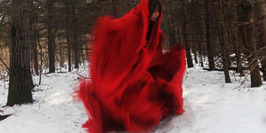 A photo shows a woman in movement, wearing a red blanket-like dress and surrounded by trees with snow on the ground.